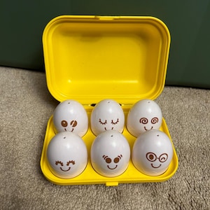 May include: A yellow plastic egg carton containing six white plastic eggs. Each egg has a different brown cartoon face drawn on it.