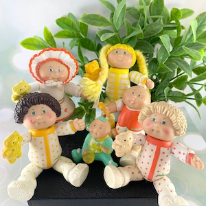 Op de afbeelding: Een groep van zes Cabbage Patch Kids poppen in verschillende outfits en poses. De poppen dragen pyjama's met stippen en strepen. Sommige poppen houden knuffels vast. De poppen zitten op een zwarte doos.