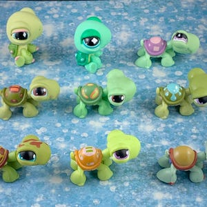 May include: A collection of ten green, blue and purple plastic turtle figurines with different coloured shells and eyes. The turtles are all in various poses, some with their heads up and some with their heads down.