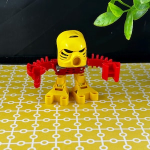 May include: A yellow and red action figure with a mask-like head, red arms, and yellow legs. The figure has a distinct design with a series of ridges and a circular opening on the head. The figure is standing on a yellow patterned surface.