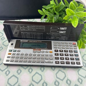 May include: A Sharp PC-1460 pocket computer with a silver keyboard, a black display frame, and a hinged lid featuring printed technical instructions. The device includes a digital screen showing the number 1234567890 and a full QWERTY layout.