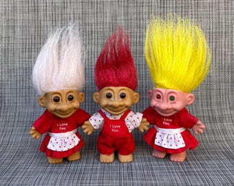 May include: Three troll dolls with tall, vibrant hair in white, red, and yellow. Each doll wears a red outfit with white, heart-patterned accents and the text I Love You printed on the front. The dolls have tan skin and large eyes.
