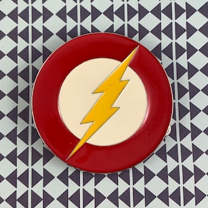 May include: A circular pin badge featuring a red border, a white central circle, and a yellow lightning bolt. The badge has a silver edge and is set against a patterned backdrop. The design is reminiscent of a superhero logo.