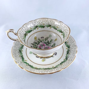 K&ouml;nnte beinhalten: Eine wei&szlig;e Teetasse und Untertasse mit einem gr&uuml;nen und goldenen Blumendesign. Tasse und Untertasse haben einen gewellten Rand.