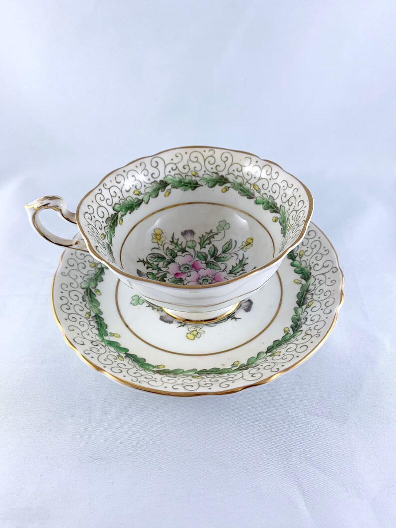 K&ouml;nnte beinhalten: Eine wei&szlig;e Teetasse und Untertasse mit einem gr&uuml;nen und goldenen Blumendesign. Tasse und Untertasse haben einen gewellten Rand.