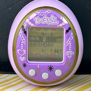 May include: A purple Tamagotchi Friends digital pet toy with a gold trim. The screen displays the time and the word "BIRTHDAY". The device has three white buttons and decorative hearts, stars, and a diamond.