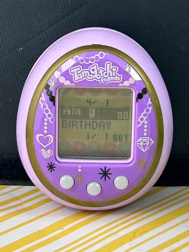 May include: A purple Tamagotchi Friends digital pet toy with a gold trim. The screen displays the time and the word "BIRTHDAY". The device has three white buttons and decorative hearts, stars, and a diamond.