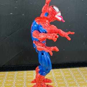 Peut inclure: Figurine articul&eacute;e de Spider-Man rouge et bleu, avec une t&ecirc;te &agrave; bouche ouverte et dents blanches, et une longue langue rose. La figurine est dans une pose dynamique, bras tendus et jambes pli&eacute;es.
