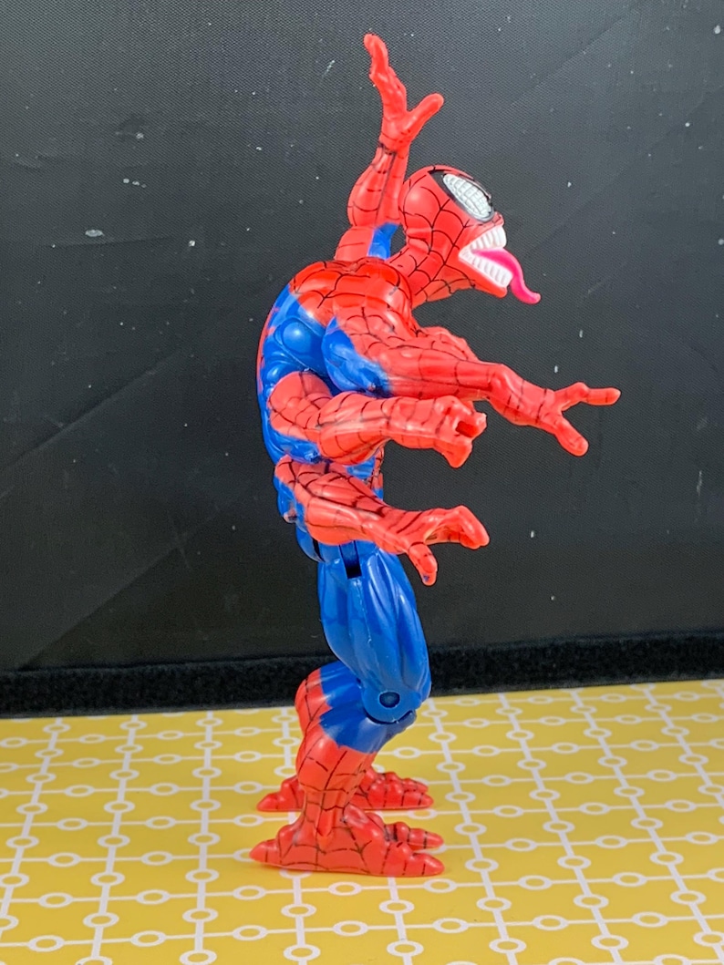 Peut inclure: Figurine articul&eacute;e de Spider-Man rouge et bleu, avec une t&ecirc;te &agrave; bouche ouverte et dents blanches, et une longue langue rose. La figurine est dans une pose dynamique, bras tendus et jambes pli&eacute;es.