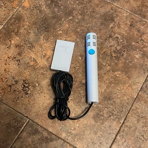 Könnte beinhalten: Ein weißes Mikrofon mit einem blauen Knopf und einem schwarzen Kabel. Das Mikrofon ist mit einem weißen Adapter verbunden, auf dem "Nintendo GameCube" steht.