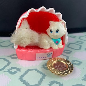 May include: A vintage toy set featuring a white cat figurine with blue eyes and a teal collar, resting in a pink bed with a red interior. A small gold-coloured plate with a brown centre is also present.