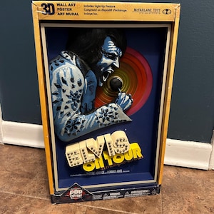 May include: A 3D wall art poster of Elvis Presley on tour. The poster features a light-up feature and is made by McFarlane Toys. The poster is in a blue and yellow box with the words "Elvis on Tour" in large white letters.