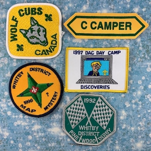 Peut inclure: Cinq écussons brodés avec différents motifs. Un écusson jaune avec un loup et le texte "CUBS WOLF CANADA". Un écusson jaune avec le texte "C CAMPER". Un écusson jaune avec une étoile verte et le texte "WHITBY DISTRICT 1996 MAP MYSTERY". Un écusson jaune avec le texte "1997 DAC DAY CAMP DISCOVERIES 1992". Un écusson vert avec deux drapeaux à damier et le texte "WHITBY DISTRICT PINE WOOD DERBY".