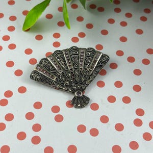 May include: A vintage silver-toned fan-shaped brooch with marcasite detailing and small, rectangular pink gemstones. The brooch has an ornate design and a small, round pink gemstone at the base. The brooch is set against a white background with red polka dots.
