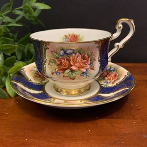 May include: A vintage teacup and saucer set featuring a floral design. The cup and saucer are white with gold trim and a blue and gold pattern. The floral design includes pink, red, and yellow roses.