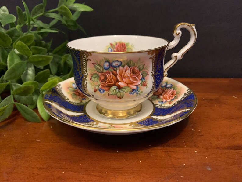 May include: A vintage teacup and saucer set featuring a floral design. The cup and saucer are white with gold trim and a blue and gold pattern. The floral design includes pink, red, and yellow roses.