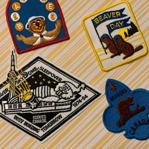 Peut inclure: Plusieurs écussons brodés vintage sur le thème des castors et du scoutisme. Les écussons comprennent un arc bleu avec un castor jonglant avec des lettres, un carré jaune avec des castors et un drapeau, et un écusson noir et blanc avec du texte.