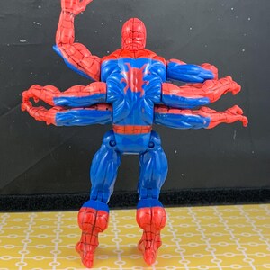 Peut inclure: Figurine articul&eacute;e de Spider-Man avec six bras, en position dynamique. La figurine est principalement bleue et rouge, avec un embl&egrave;me d'araign&eacute;e dans le dos. Elle est pos&eacute;e sur une surface jaune.