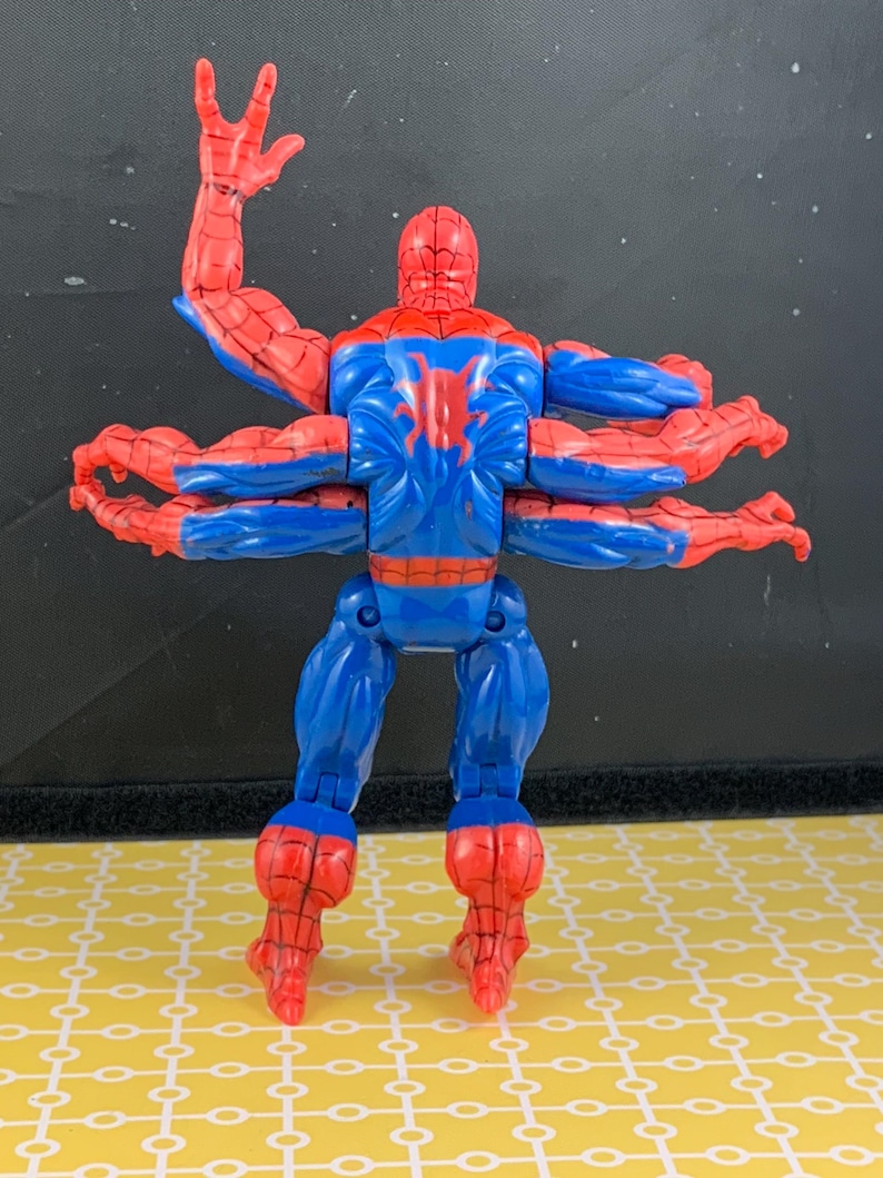 Peut inclure: Figurine articul&eacute;e de Spider-Man avec six bras, en position dynamique. La figurine est principalement bleue et rouge, avec un embl&egrave;me d'araign&eacute;e dans le dos. Elle est pos&eacute;e sur une surface jaune.