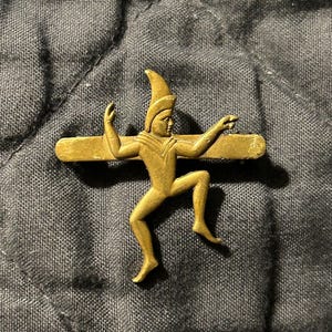 Peut inclure: Une épingle en métal dorée représentant une figure stylisée d'une personne portant un chapeau pointu, une jambe levée et les bras tendus. La figure est fixée à une barre horizontale.