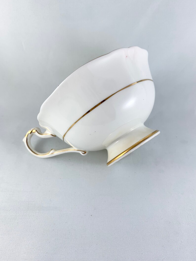 K&ouml;nnte beinhalten: Eine wei&szlig;e Teetasse mit goldener Verzierung am Rand und am Boden. Die Teetasse hat einen zarten Henkel und einen gewellten Rand.