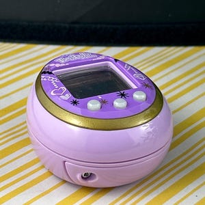 May include: A lavender Tamagotchi virtual pet device with a gold trim and three white buttons. The screen is black, and the device has a small speaker and a port. The top of the device has the text "Tamagotchi" in a decorative font.