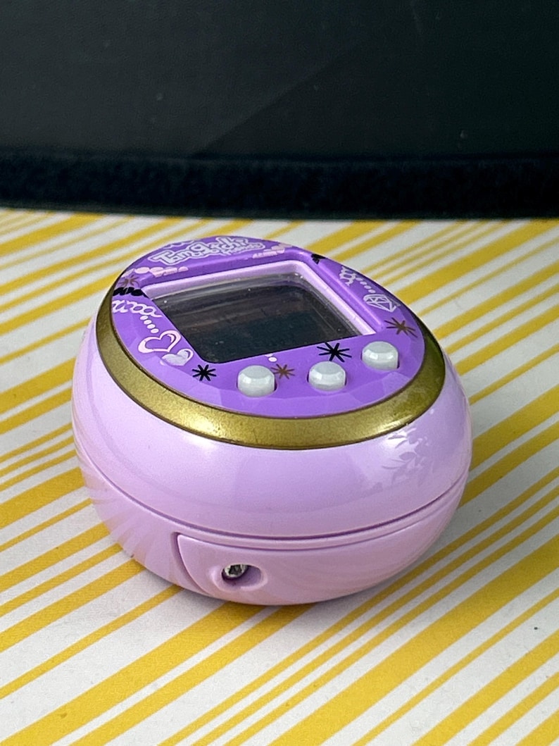 May include: A lavender Tamagotchi virtual pet device with a gold trim and three white buttons. The screen is black, and the device has a small speaker and a port. The top of the device has the text "Tamagotchi" in a decorative font.