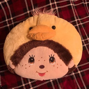 Peut inclure: Un coussin rond en peluche avec une tête de canard jaune et un visage aux cheveux bruns. Le canard a un bec orange et des yeux noirs. Le visage a des yeux brodés, des taches de rousseur et une bouche rouge souriante. Le coussin est sur une couverture à carreaux rouge et noir.