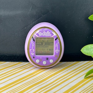 May include: A purple Tamagotchi Friends digital pet toy. The egg-shaped device has a pink border with gold accents and a small screen displaying numbers and the word "BIRTHDAY". The device has three white buttons and is decorated with hearts, stars, and a diamond.