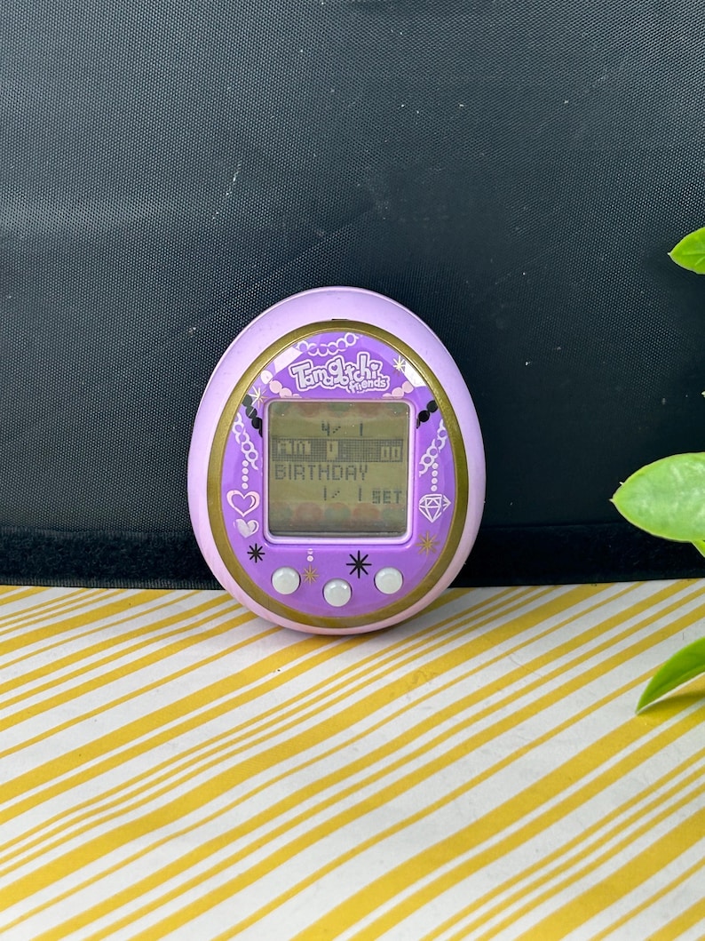 May include: A purple Tamagotchi Friends digital pet toy. The egg-shaped device has a pink border with gold accents and a small screen displaying numbers and the word "BIRTHDAY". The device has three white buttons and is decorated with hearts, stars, and a diamond.