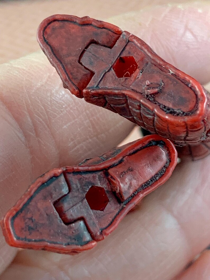 May include: Two red, textured, snake head pieces with a dark border. Each piece has a hexagonal hole and a connecting notch. The pieces are held in a hand.