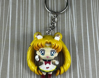 Op de afbeelding: Een gele Sailor Moon sleutelhanger met een miniatuurfiguur met een witte jurk, rode strik en laarzen. De sleutelhanger heeft een zilveren ketting en ring. Het figuur heeft grote blauwe ogen en geel haar. De achtergrond is gestreept.