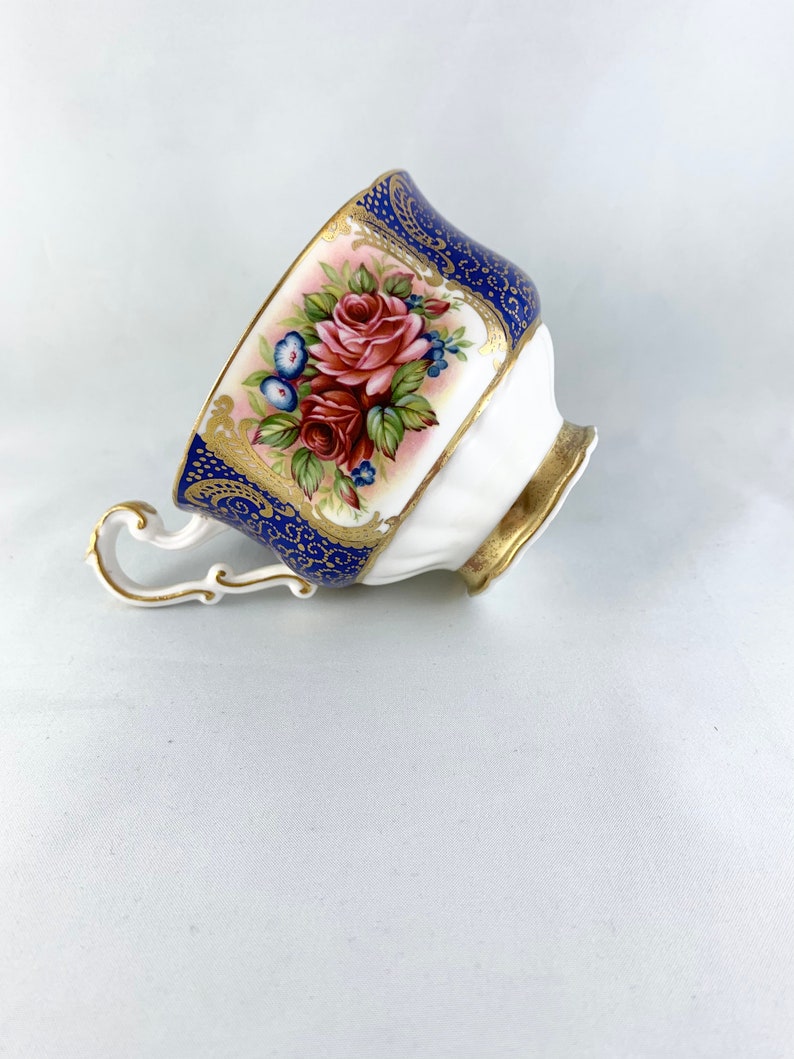 May include: A white teacup with a blue and gold floral design. The cup has a delicate handle and a scalloped rim.