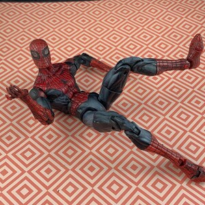 May include: A Spider-Man action figure in a dynamic pose, featuring a red and black costume with web detailing. The figure is lying on a patterned surface with a repeating geometric design.