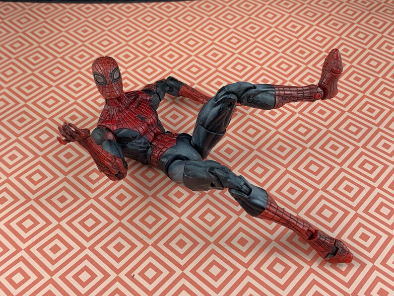 May include: A Spider-Man action figure in a dynamic pose, featuring a red and black costume with web detailing. The figure is lying on a patterned surface with a repeating geometric design.