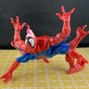 Peut inclure: Figurine articul&eacute;e de Spider-Man, en rouge et bleu, avec un embl&egrave;me d'araign&eacute;e blanc sur la poitrine. La figurine a plusieurs bras et une langue rose. Elle est pos&eacute;e sur une surface jaune &agrave; motifs.