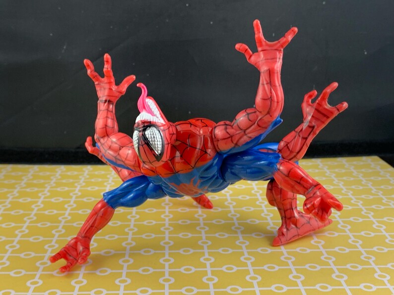 Peut inclure: Figurine articul&eacute;e de Spider-Man, en rouge et bleu, avec un embl&egrave;me d'araign&eacute;e blanc sur la poitrine. La figurine a plusieurs bras et une langue rose. Elle est pos&eacute;e sur une surface jaune &agrave; motifs.