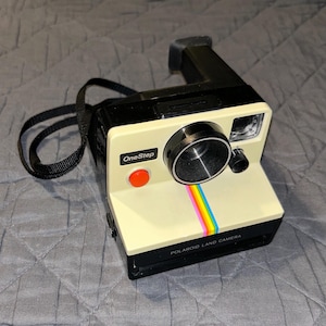 Peut inclure: Un appareil photo Polaroid OneStep Land vintage beige avec un objectif noir et une bande arc-en-ciel. L'appareil photo est doté d'une sangle noire et d'un bouton rouge portant l'inscription "OneStep".