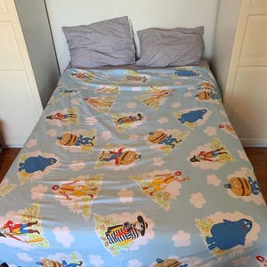 May include: A light blue bedsheet with a repeating pattern of cartoon characters, including a hamburger, a cowboy, and a blue creature. The sheet is on a bed with two gray pillows. The bed has a black and white patterned border.