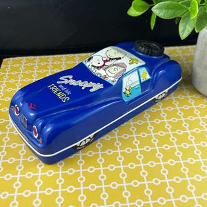 May include: A blue Snoopy and Friends toy car with white trim. The car features Snoopy and Woodstock illustrations on the roof and side windows. The text "Snoopy and his Friends" is displayed on the side of the car.