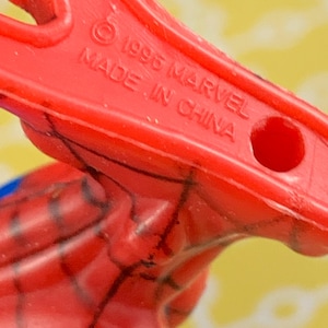 Peut inclure: Gros plan sur une pi&egrave;ce de jouet en plastique rouge avec le texte "&copy; 1996 MARVEL MADE IN CHINA". La pi&egrave;ce de jouet a un trou et fait partie d'un jouet plus grand, avec des lignes noires visibles.