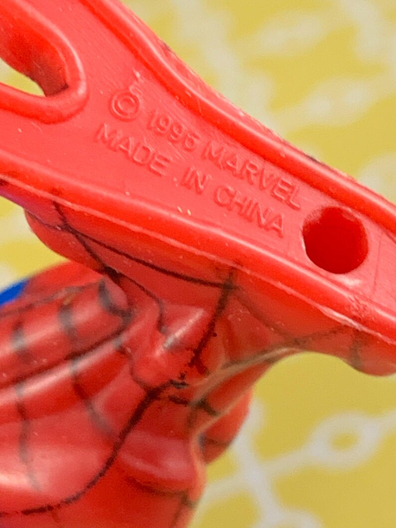Peut inclure: Gros plan sur une pi&egrave;ce de jouet en plastique rouge avec le texte "&copy; 1996 MARVEL MADE IN CHINA". La pi&egrave;ce de jouet a un trou et fait partie d'un jouet plus grand, avec des lignes noires visibles.