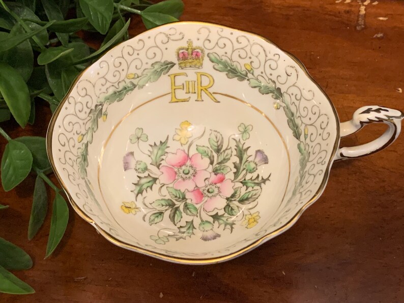 K&ouml;nnte beinhalten: Eine wei&szlig;e Teetasse mit goldfarbenem Rand und floralem Design. Die Tasse zeigt eine goldene Krone und die Buchstaben "E II R" in der Mitte.