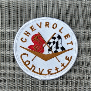 May include: A circular embroidered patch with a white background and a white stitched border. The design features the brown text CHEVROLET at the top and CORVETTE at the bottom. In the center are two crossed flags, one red with a gold fleur-de-lis and one black and white checkered, positioned above a gold V-shaped emblem.