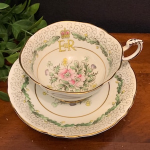 K&ouml;nnte beinhalten: Eine wei&szlig;e Teetasse und Untertasse mit goldener Verzierung und Blumenmuster. Die Tasse zeigt eine Krone und die Buchstaben "ER" in Gold. Die Untertasse hat einen gr&uuml;nen und goldenen Blumenrand.