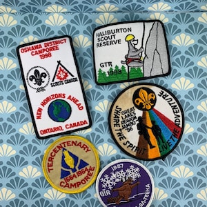 Peut inclure: Une collection de neuf écussons de scout brodés. Les écussons sont de différentes couleurs et présentent différents motifs, notamment une tente, un castor, un globe terrestre et un alpiniste. Les écussons proviennent tous de différents camps et événements scouts.
