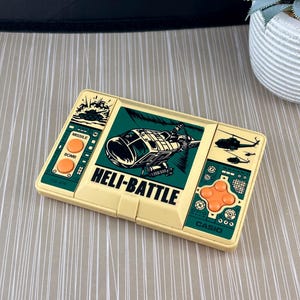 May include: A vintage Casio handheld game called "Heli-Battle" with a green and brown colour scheme. The game features a helicopter and a tank on the screen. The game has buttons for missile and bomb attacks.