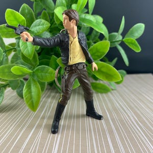 May include: A miniature figurine of a character from a science fiction film, dressed in a black jacket, tan shirt, and brown pants, holding a blaster. The figure is posed with the blaster extended, as if in action. The figure is approximately 3 inches tall.