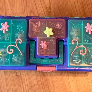 May include: A blue and purple plastic toy case with multiple compartments. The case has a pink clasp and is decorated with pink and yellow flowers, swirls, and small animal figures. The compartments are clear and allow visibility of the contents.