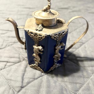 May include: A small blue and silver teapot with a rabbit finial on the lid. The teapot has a hexagonal body with silver accents and animal figures.